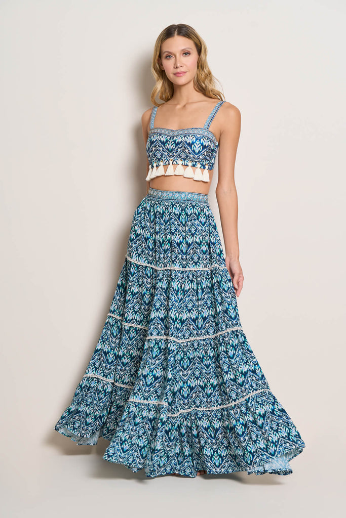 A woman casually posing, wearing the Maxi Skirt, in a blue and white pattern combination, with tiered skirt and white embroidery works to accentuate the pattern, in a full length size, paired with a bikini top.