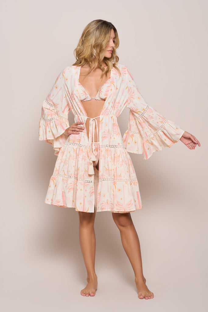 A model casually posing while wearing the eBarbie Short Kimono in white and light pink floral patterns, with embroidered patterns on the ruffles both on the bottom and on the hand flair, with a tie up belt, a perfect coverup for the same colored bikini top and bottom.